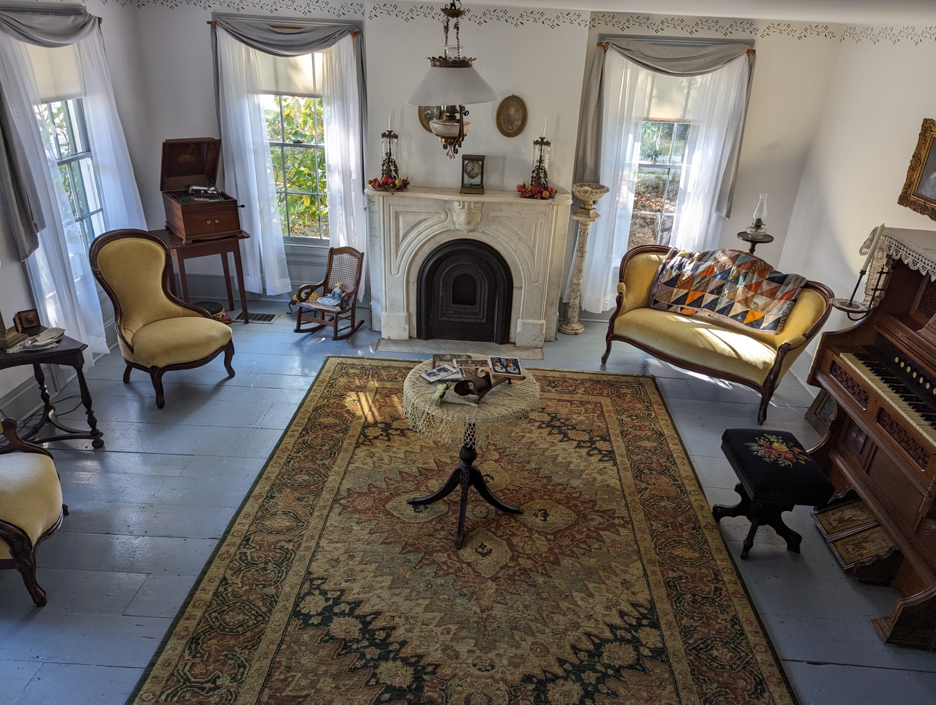 Historic Victorian parlor with a carved marble fireplace, antique furniture, upholstered settee and chairs, vintage organ and phonograph, patterned area rug, and tall sunlit windows with sheer curtains.
